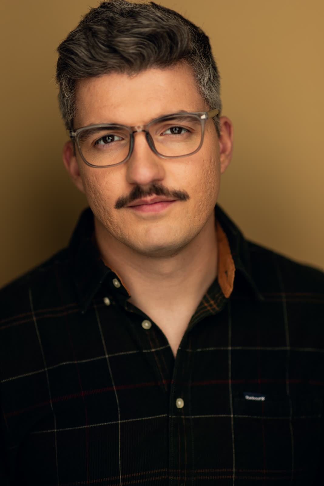 Eugene Romanchuk, theatrical headshot: glasses and dark plaid shirt against a mustard background